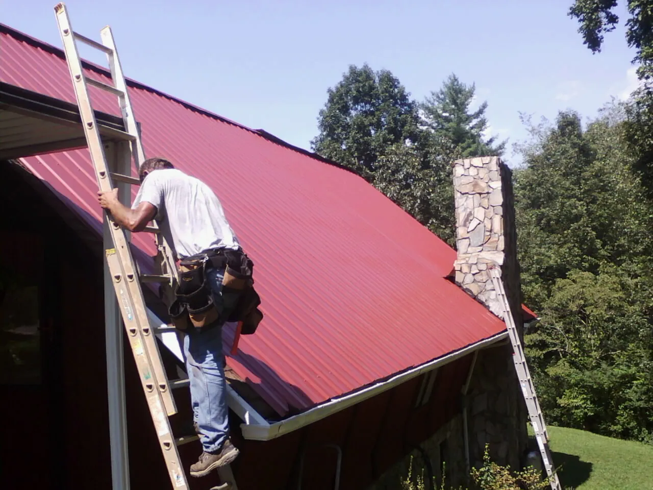 Roofer on a ladder beside a red metal roof and stone chimney — Soffit & Fascia Repair by Roofing Brothers in Rosburg